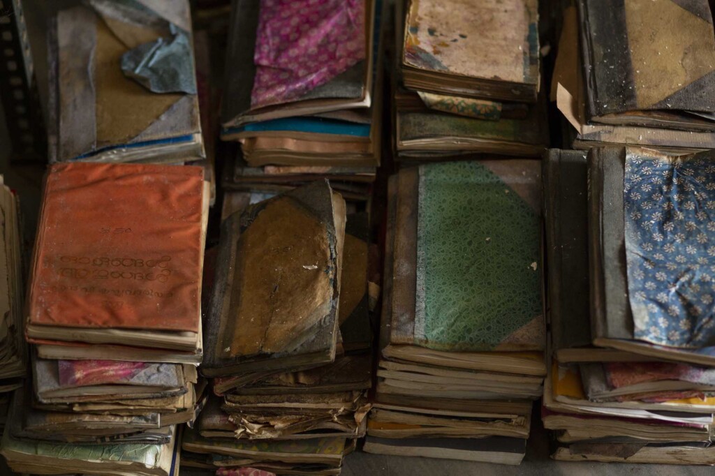 In the photograph, stacks of old, worn books can be seen. The books are arranged in two horizontal rows, covering the entire image.