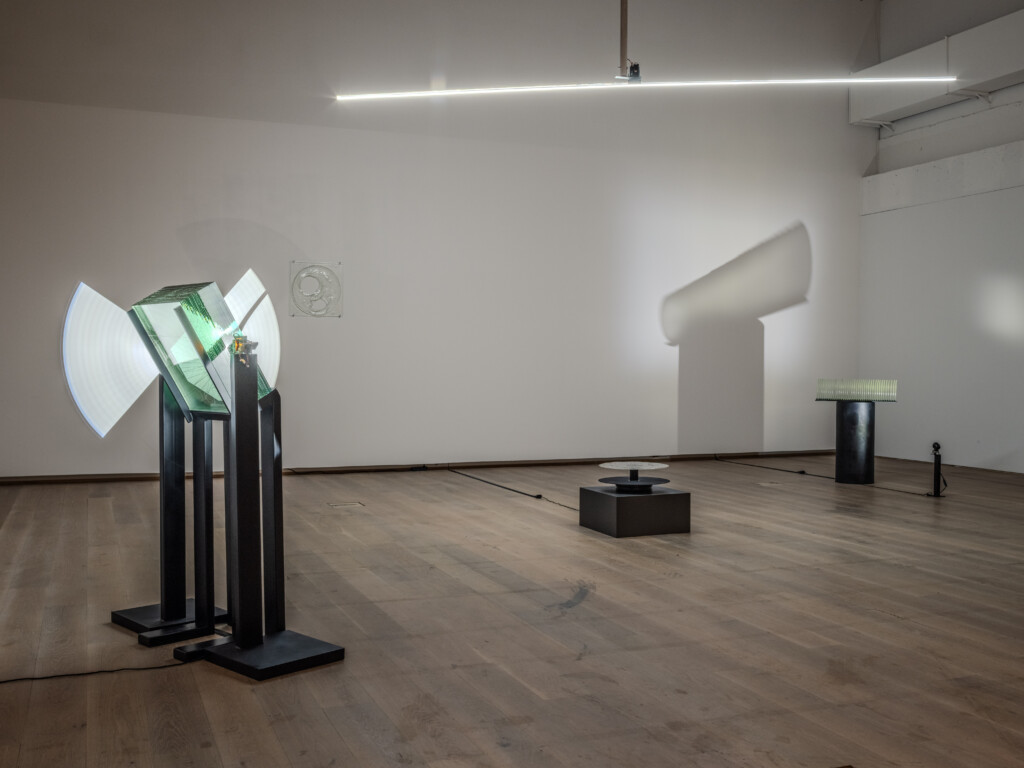 Glass sculptures illuminated by strong light sticks that create shadows along white walls.