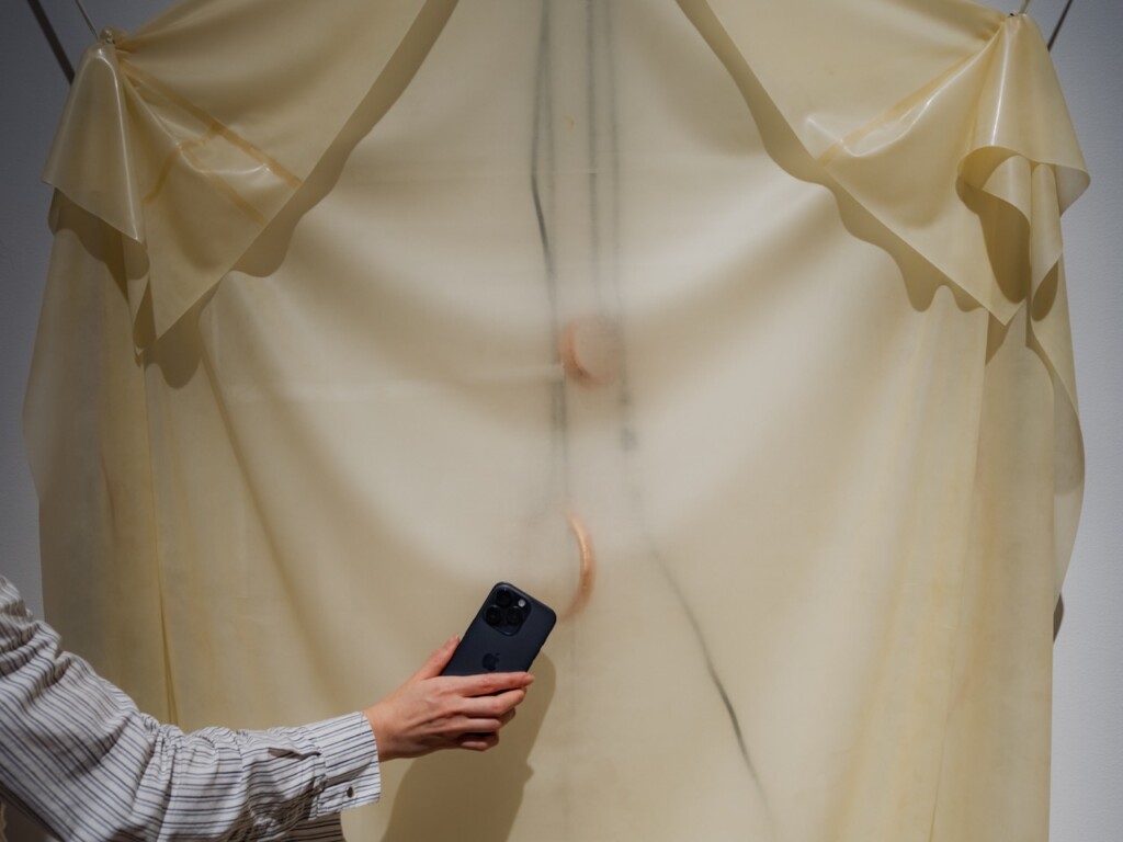 Närbild på en vägghängd böljande draperad latex -skulptur i ljusgult. En hand med en mobiltelefon svävar framför skulpturen.