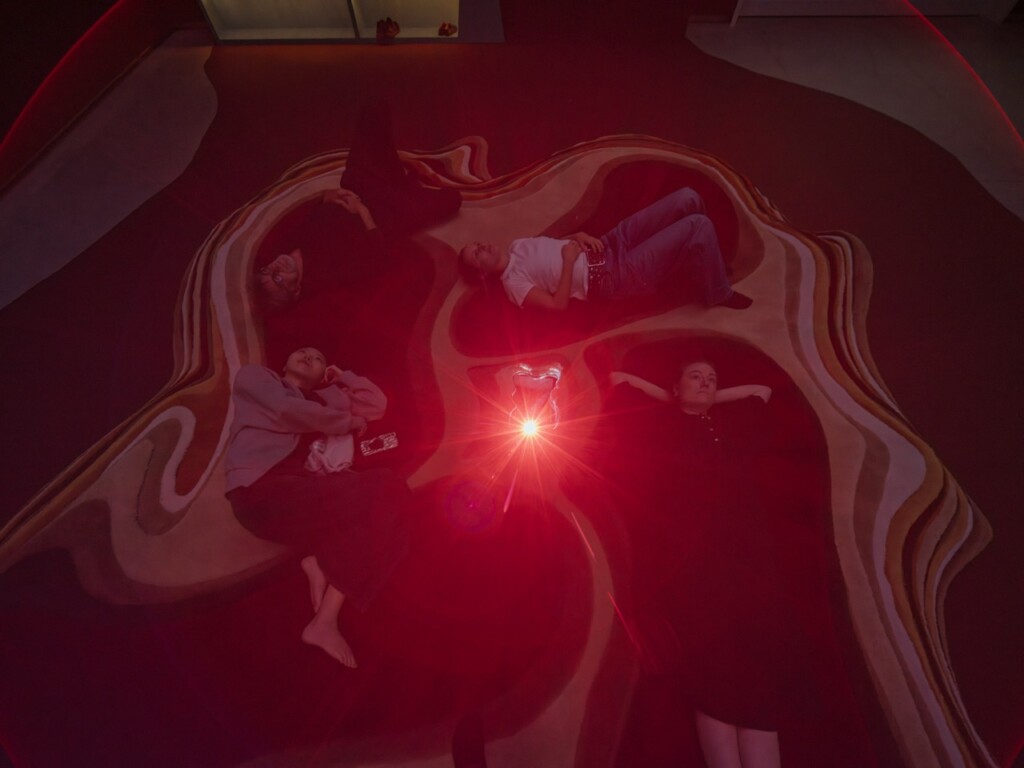 Four people lie down on a pile of rugs and look up, a red light shining in the middle of the picture.
