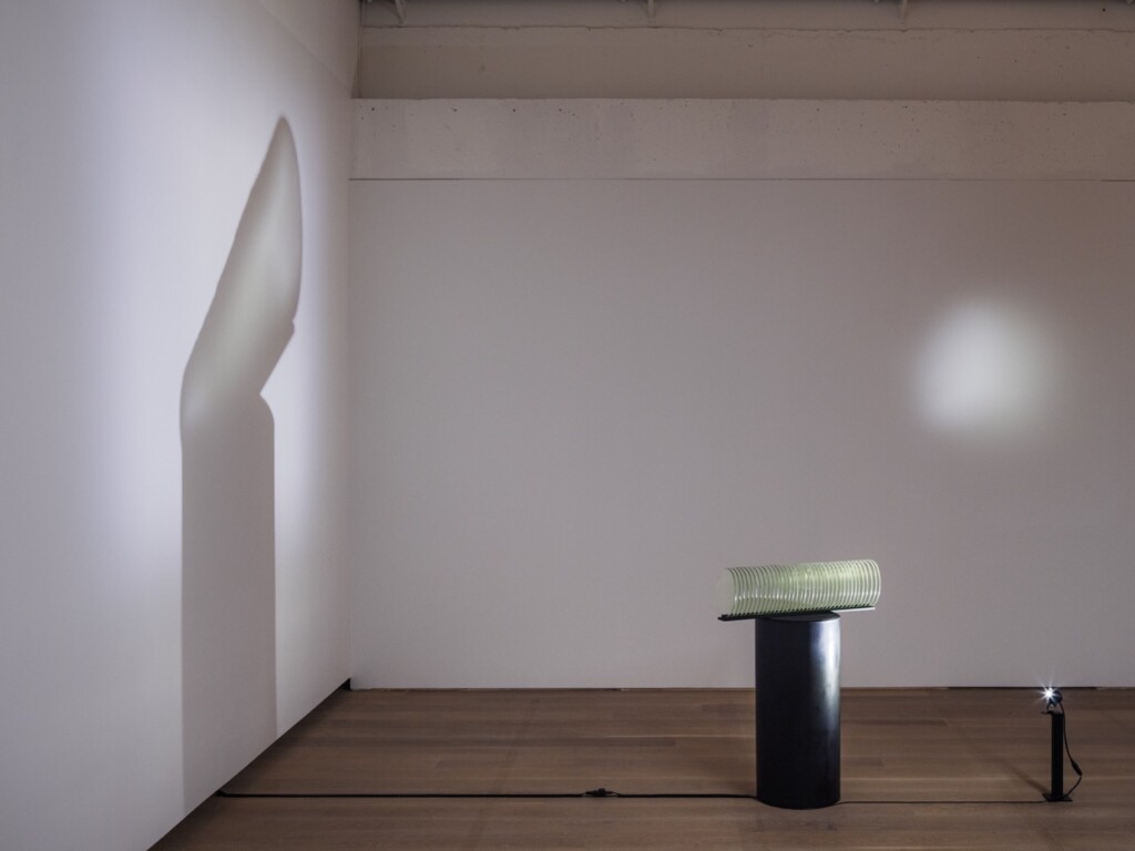Glasskulptur belysta med ljus som skapar skuggor längs vita väggar.